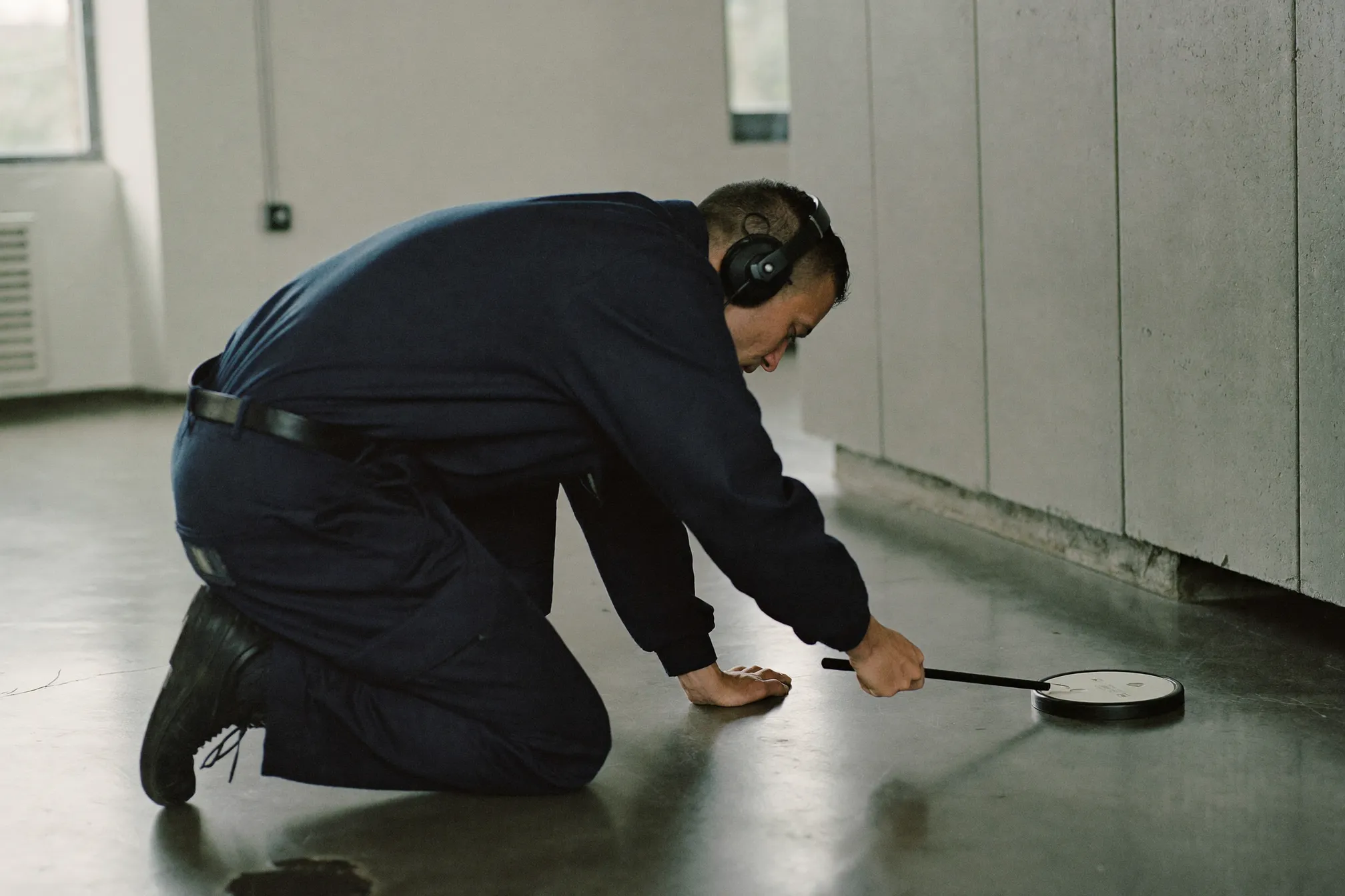 A leak-detection technician kneeling on a concrete floor, using an acoustic ground microphone and headphones to locate a water leak beneath the slab