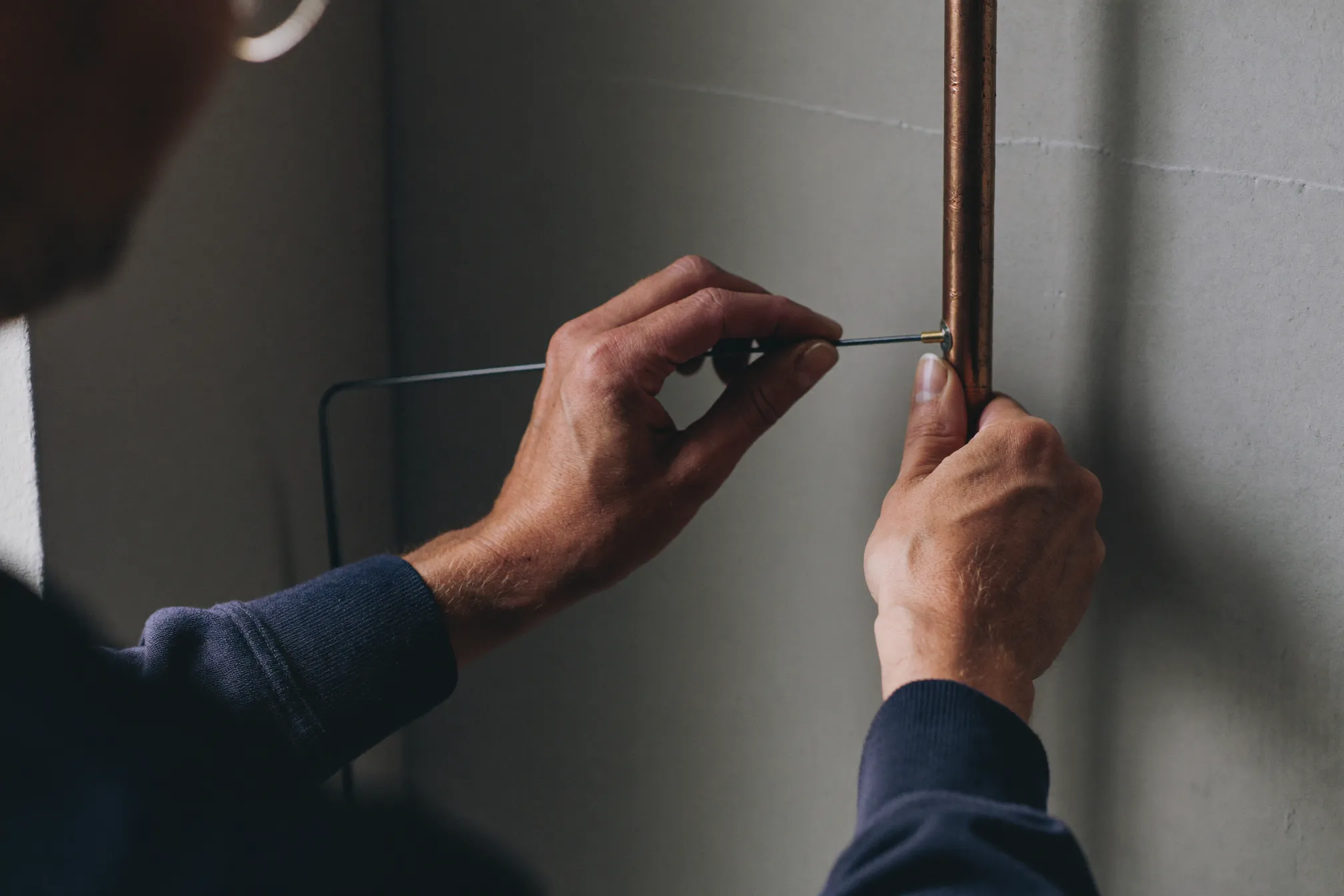 A technician's hands pressing a slim acoustic listening rod against a copper water pipe to trace a leak without opening the wall